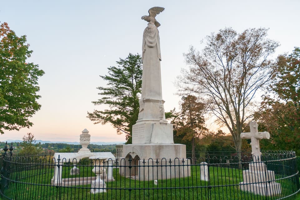 A statue of a figure with an eagle, surrounded by a cemetery fence, with trees and a cross in the background.
