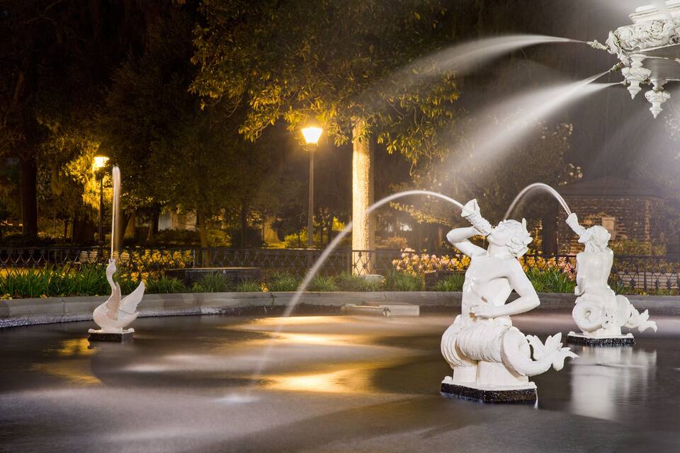 A nighttime fountain with swan and human figures, surrounded by trees and illuminated by street lamps.