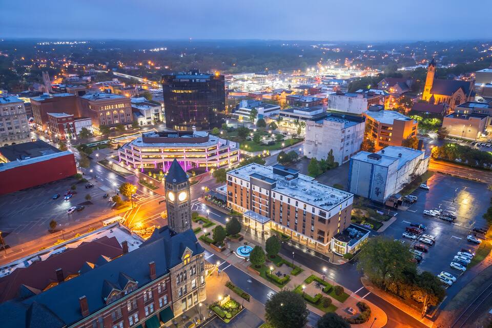 A cityscape at dusk with illuminated buildings, a prominent clock tower, and a church spire.