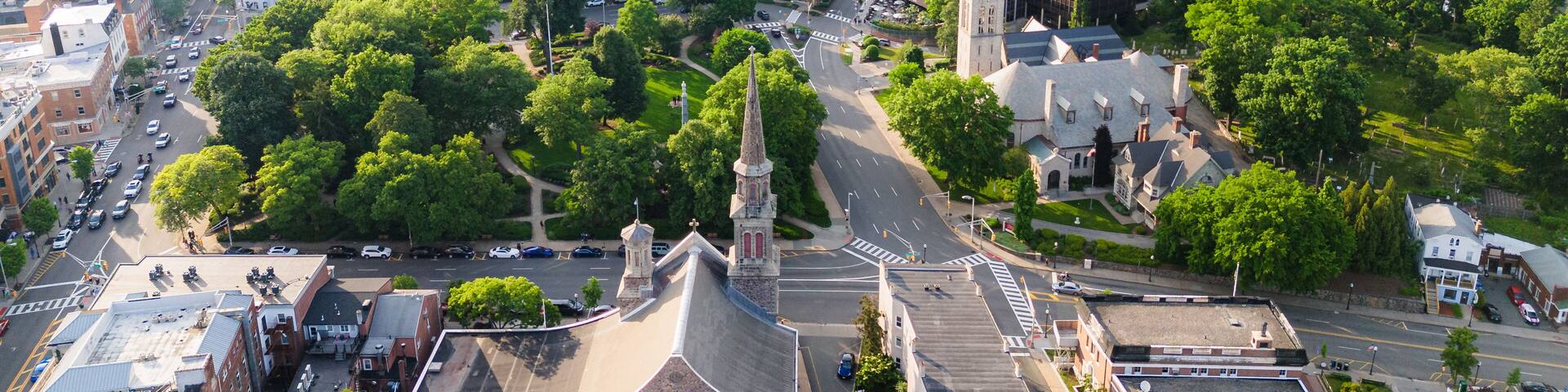 Une vue aérienne d’une ville où se côtoient des bâtiments modernes et anciens, avec un clocher d’église imposant et une rivière en arrière-plan.