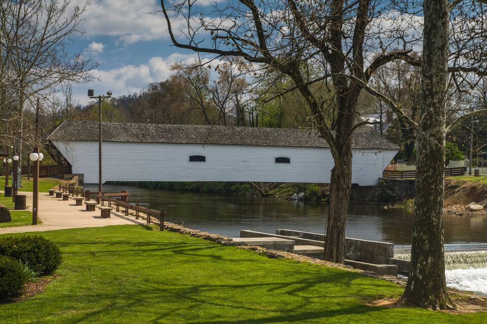 A white wooden structure by a river, surrounded by trees and a grassy area with benches.