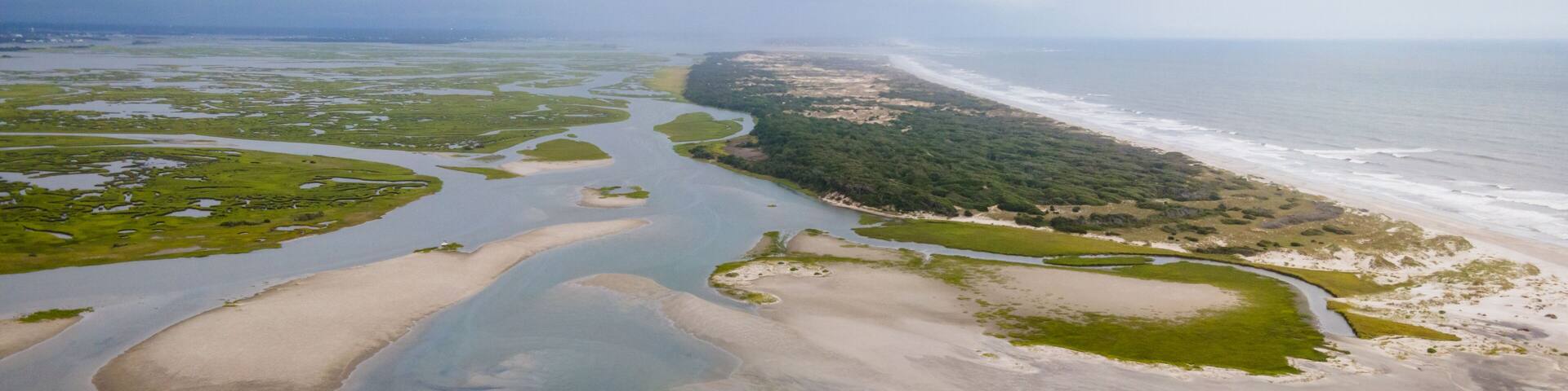 Vista aérea de un paisaje costero con un río, zonas arenosas y una península boscosa.