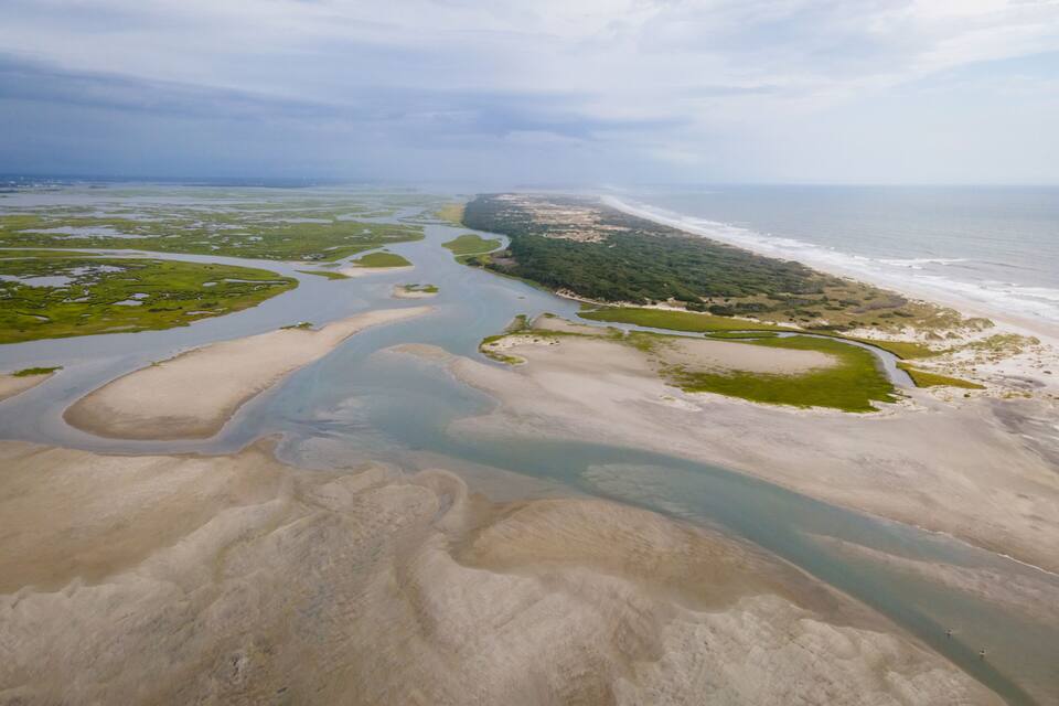 An aerial view of a coastal landscape with a river, sandy areas, and a forested peninsula.