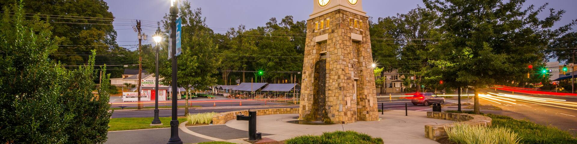 A stone clock tower with two faces, surrounded by grass and trees, at dusk with street lights on.