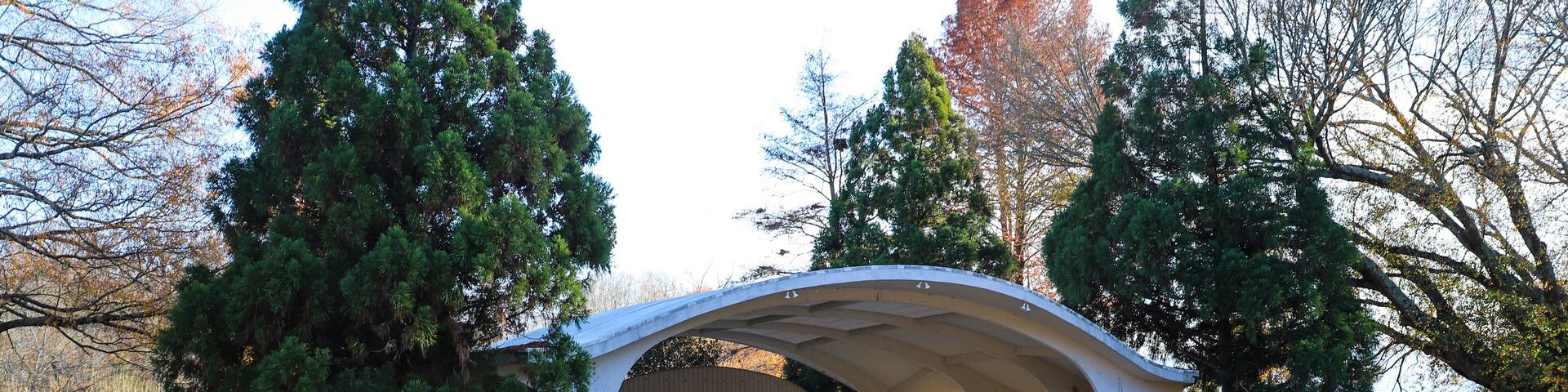 A covered pavilion with a curved roof, surrounded by trees and a stone wall.