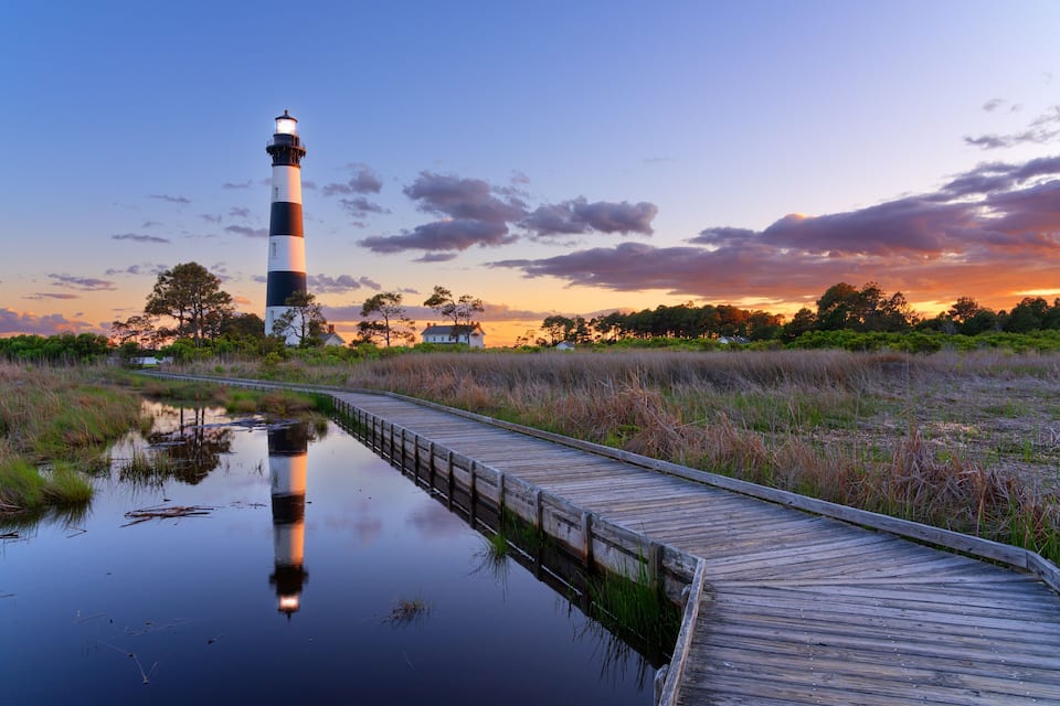 A lighthouse, a wooden boardwalk, and a sunset over a marshy landscape.