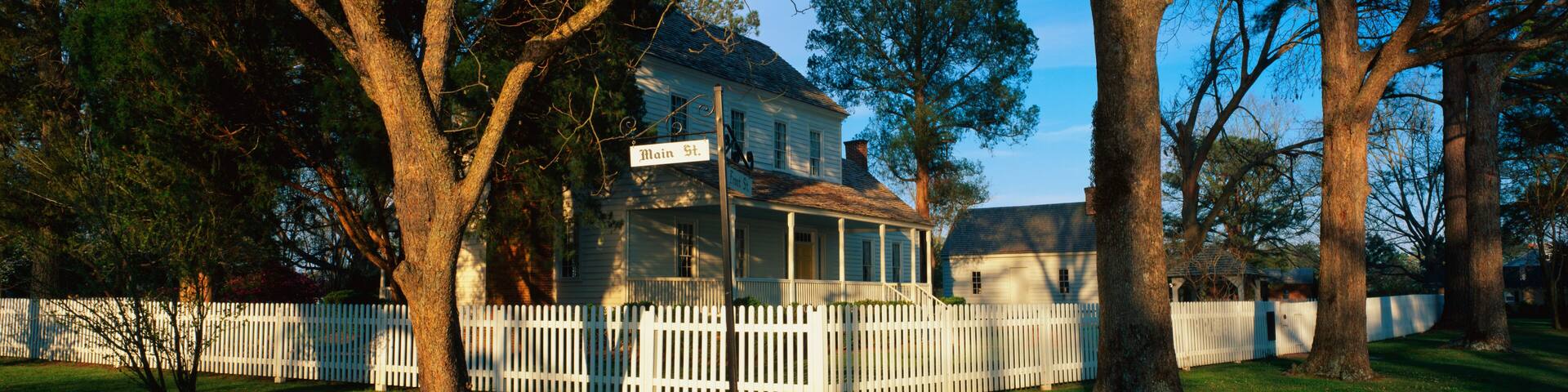 A white house with a porch, surrounded by a white picket fence, and large trees in the background.