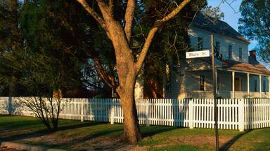 A white house with a porch, surrounded by a white picket fence, and large trees in the background.
