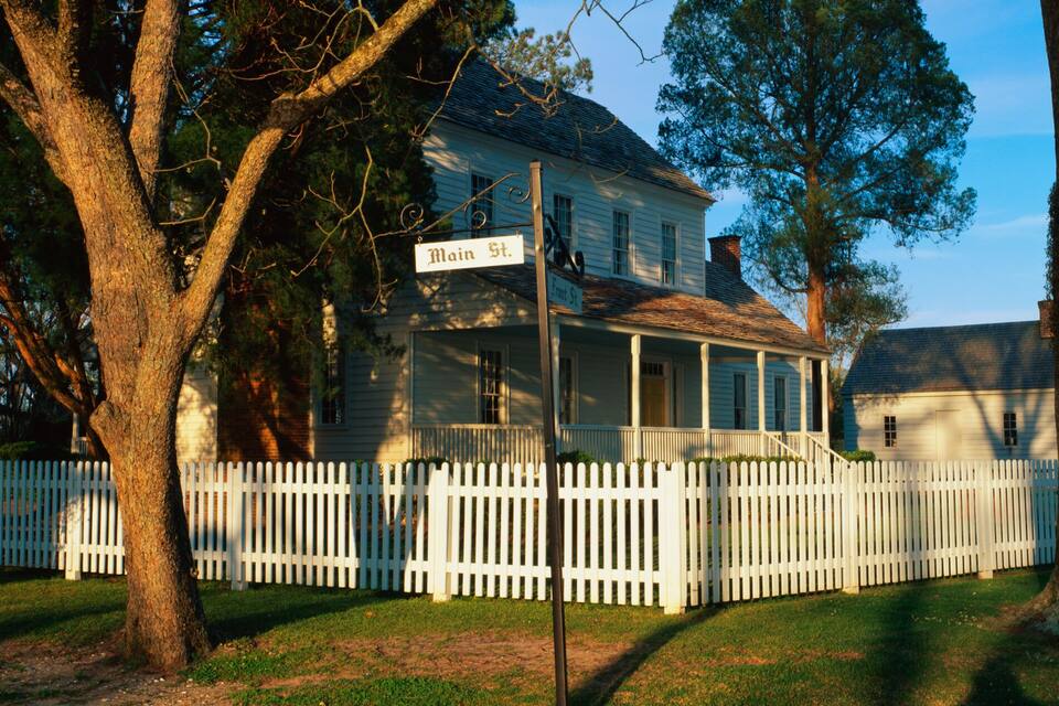 A white house with a porch, surrounded by a white picket fence, and large trees in the background.