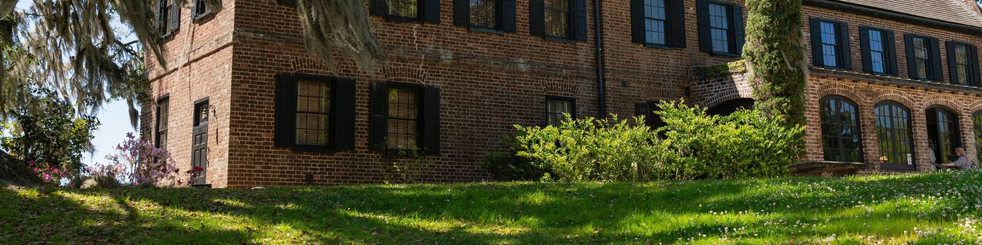 A large brick building with multiple chimneys, surrounded by lush greenery and a tall cypress tree.