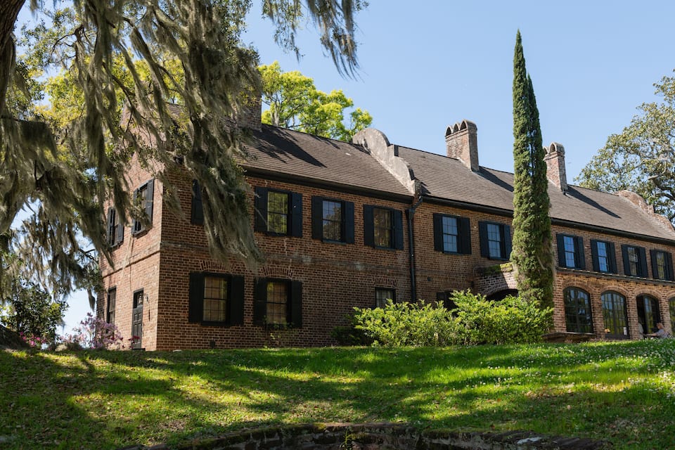 A large brick building with multiple chimneys, surrounded by lush greenery and a tall cypress tree.