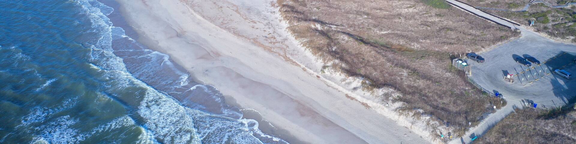 A coastal landscape with a sandy beach, a pier, and a parking area.