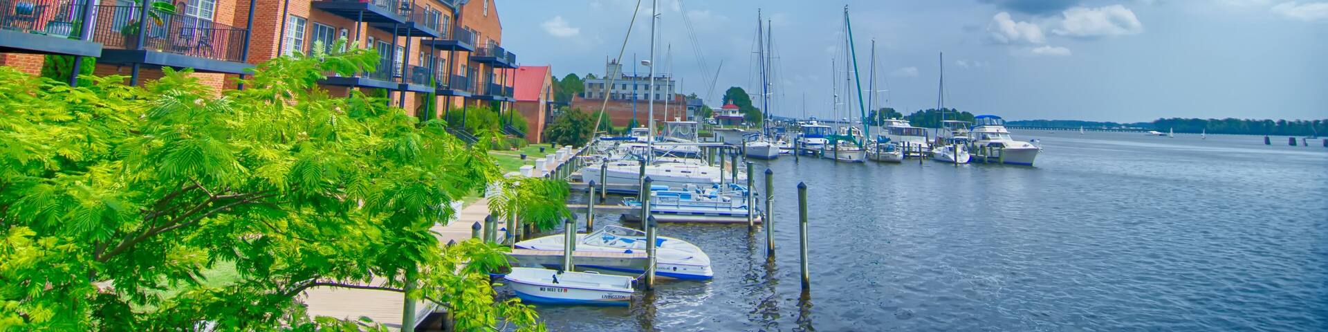 A marina with boats docked along a waterfront, surrounded by brick buildings and lush greenery.