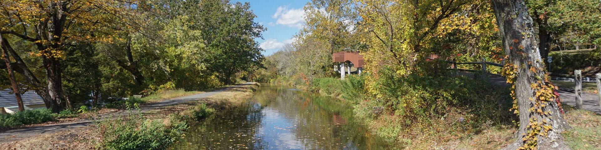 A calm river with autumn leaves, trees, and a small building in the distance.