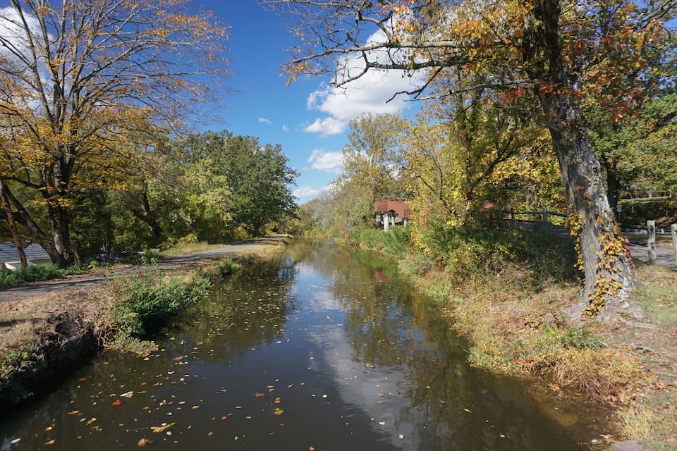 A calm river with autumn leaves, trees, and a small building in the distance.