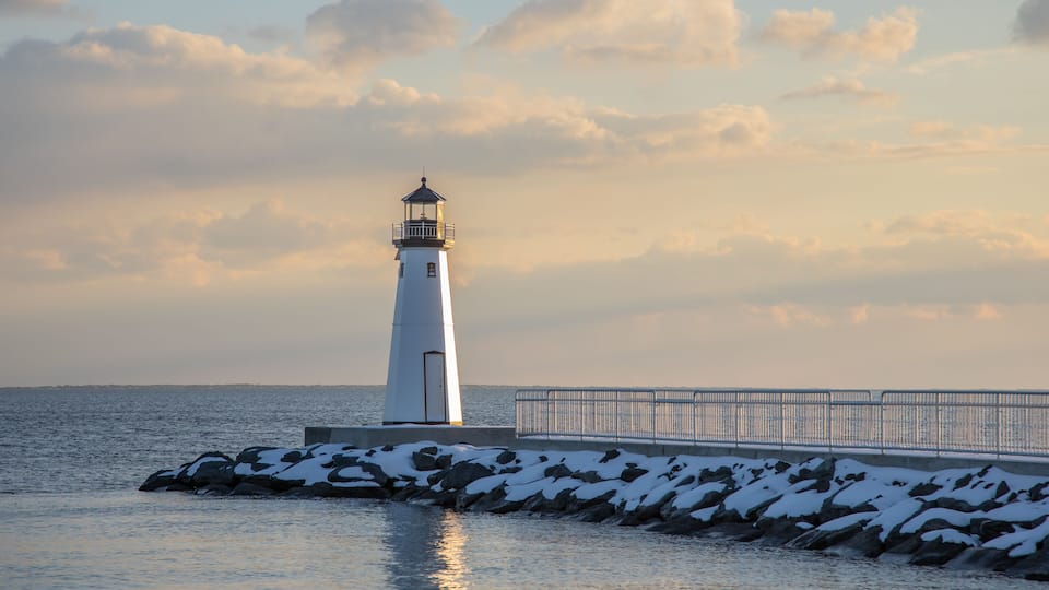 A lighthouse on a rocky pier with snow-covered rocks and a calm sea.