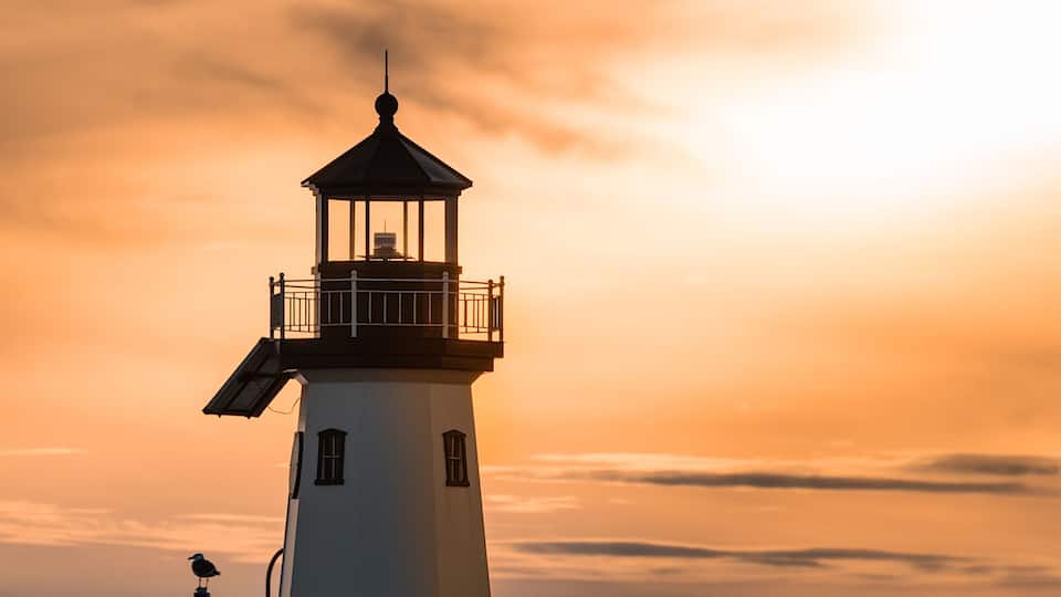 A lighthouse at sunset with a bird on top, surrounded by rocks and water.