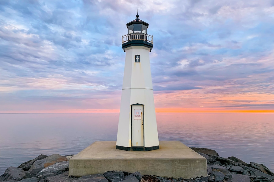 Un phare blanc, surmonté d’un panneau indiquant « DÉFENSE D’ENTRER », se dresse sur une base en béton, entouré de rochers, sur fond de ciel au coucher du soleil.