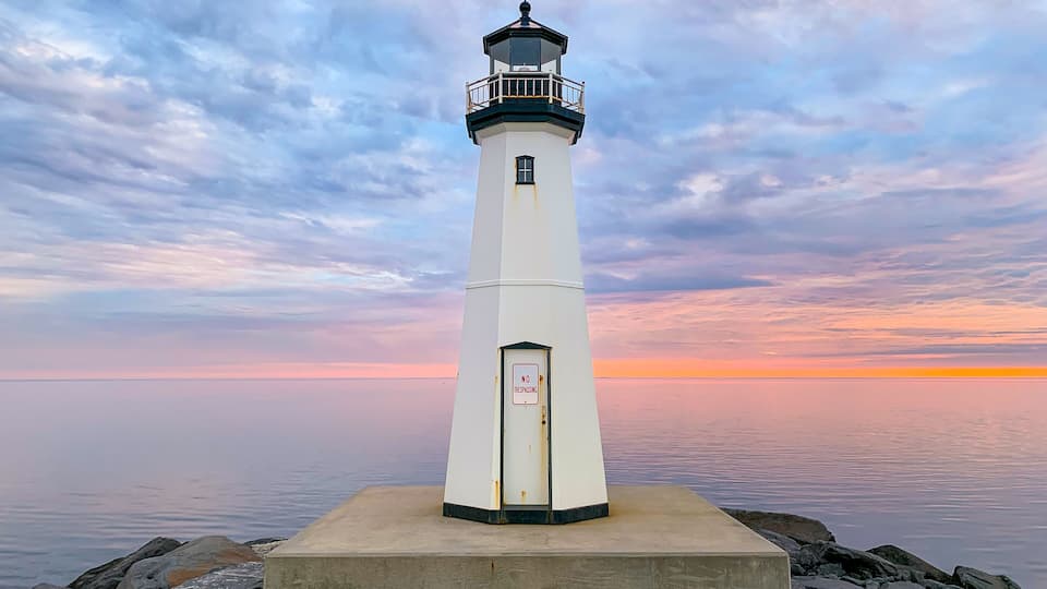 A white lighthouse with a 'NO TRESPASSING' sign, standing on a concrete base with rocks around it, against a sunset sky.