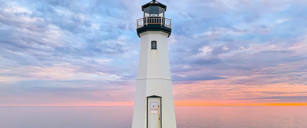 A white lighthouse with a 'NO TRESPASSING' sign, standing on a concrete base with rocks around it, against a sunset sky.