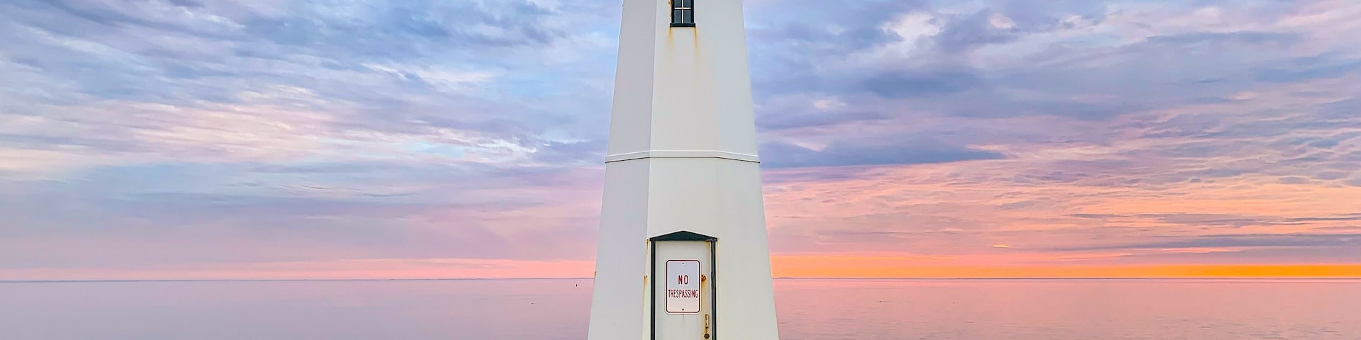 A white lighthouse with a 'NO TRESPASSING' sign, standing on a concrete base with rocks around it, against a sunset sky.