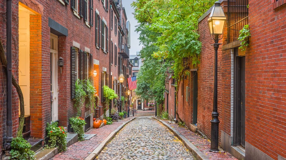 A cobblestone alley flanked by brick buildings with green shutters, a street lamp, and an American flag.