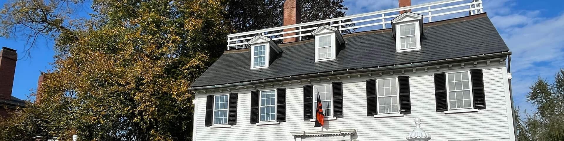 A white colonial-style house with black shutters, surrounded by a white picket fence, hay bales, and pumpkins.