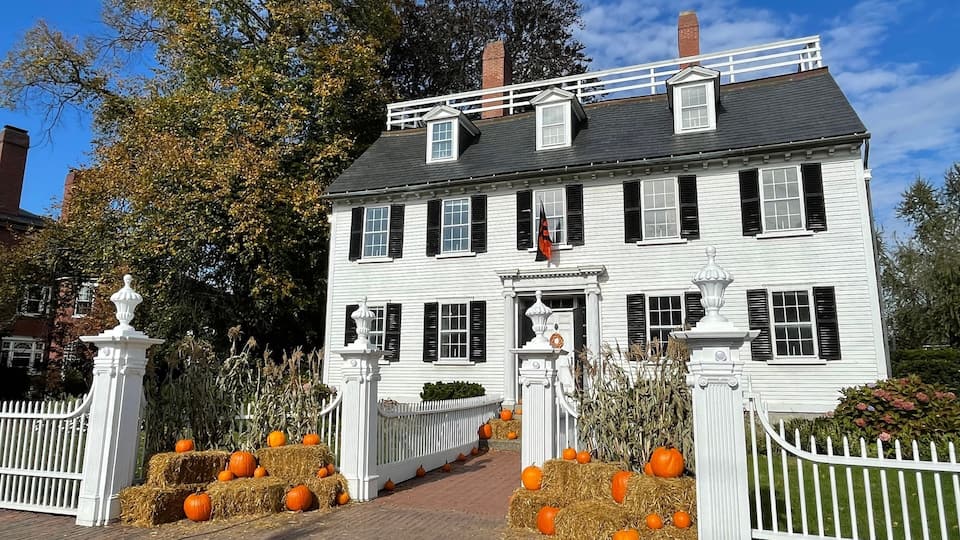 A white colonial-style house with black shutters, surrounded by a white picket fence, hay bales, and pumpkins.