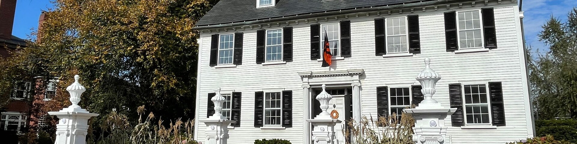 Una casa de estilo colonial blanco con contraventanas negras, rodeada por una cerca de piquetes blanca, fardos de heno y calabazas.