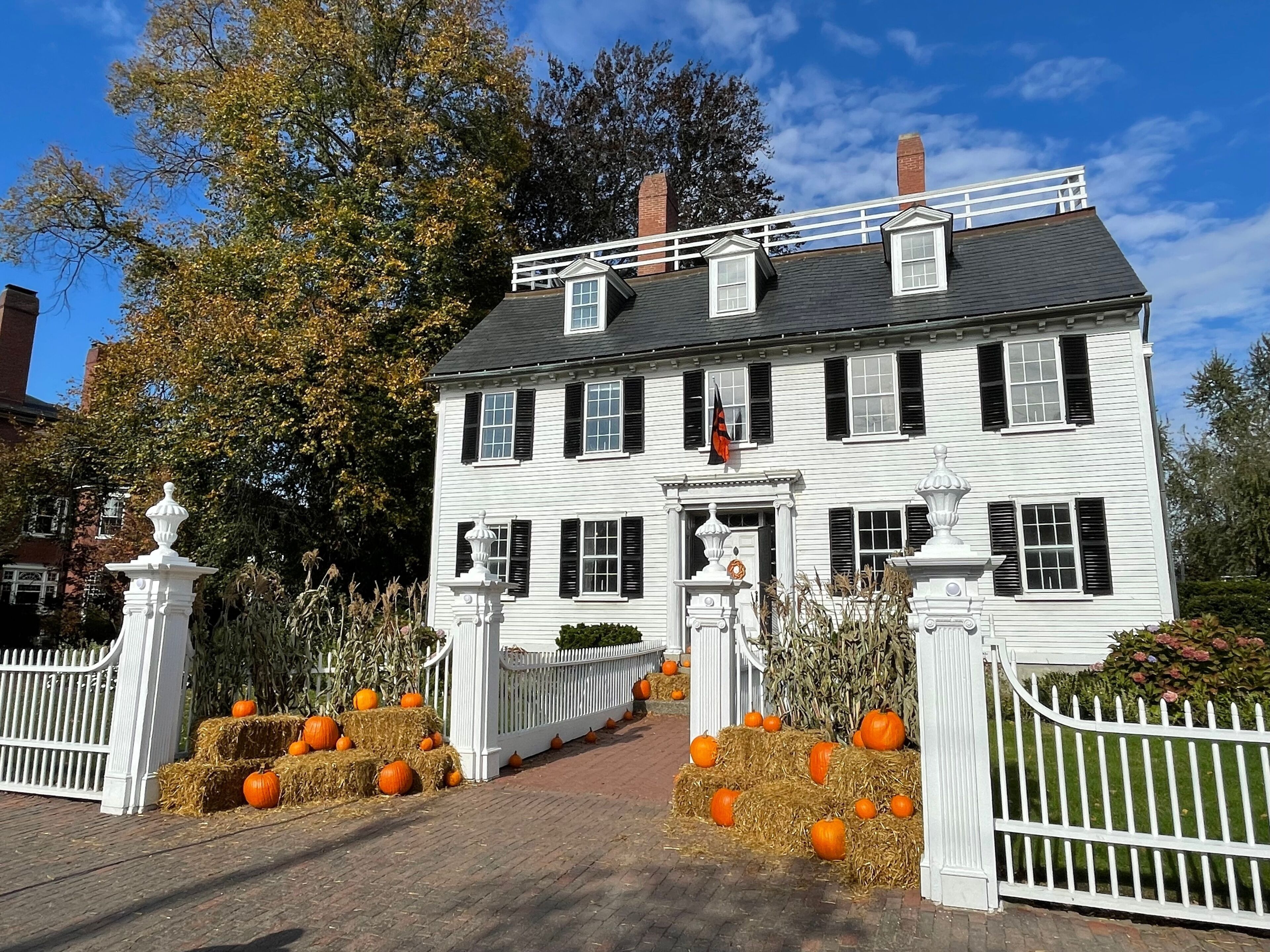 A white colonial-style house with black shutters, surrounded by a white picket fence, hay bales, and pumpkins.