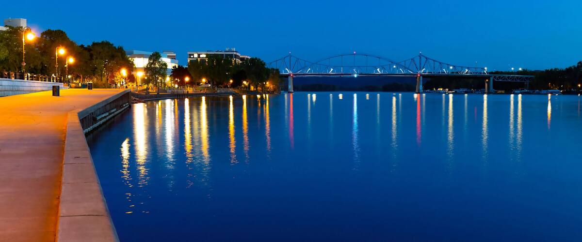 A calm river at dusk with a bridge and city lights reflecting on the water.