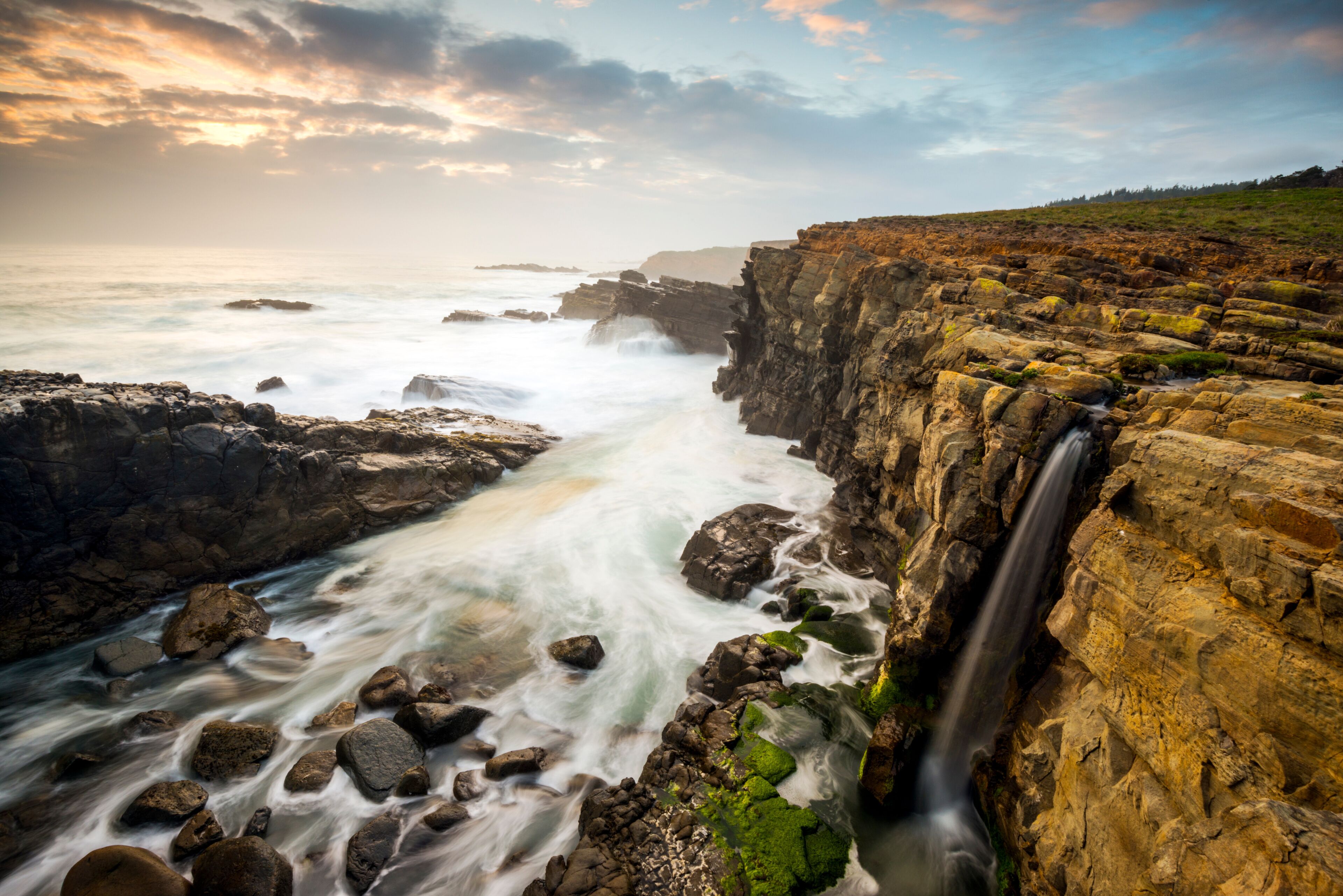 Paisagem costeira com cachoeira, terreno rochoso e um penhasco com vista para o oceano.