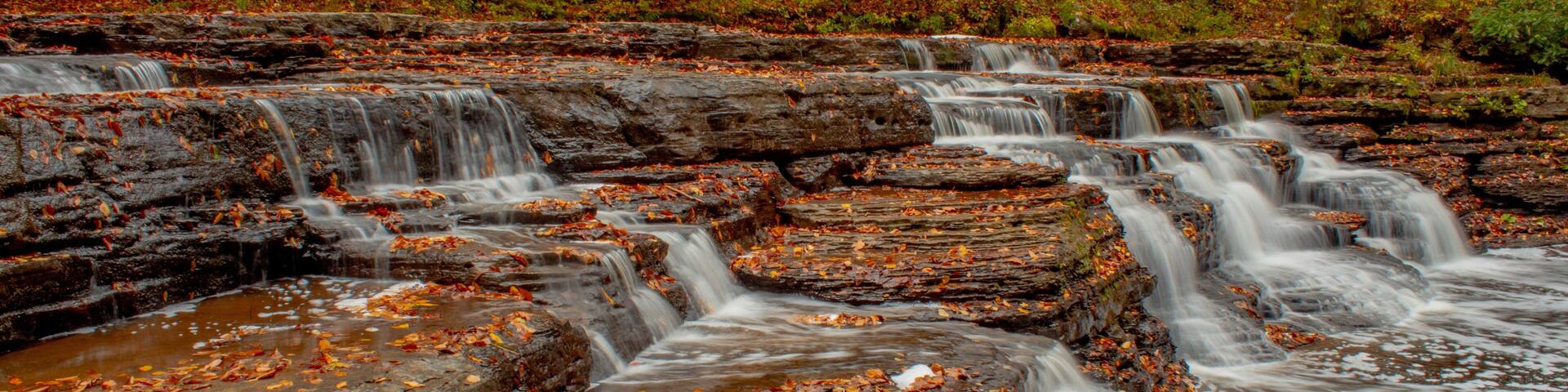 A waterfall cascading over rocky terrain with autumn leaves scattered around.