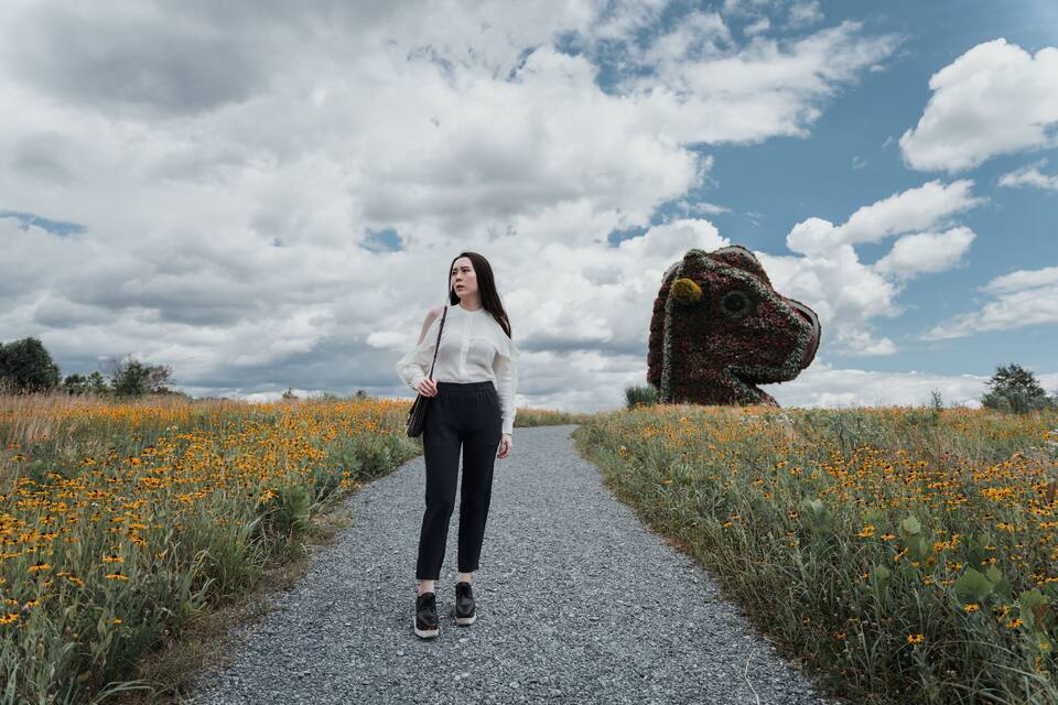 Una mujer caminando por un sendero de grava rodeada de campos de flores anaranjadas, con una gran escultura de roca al fondo.