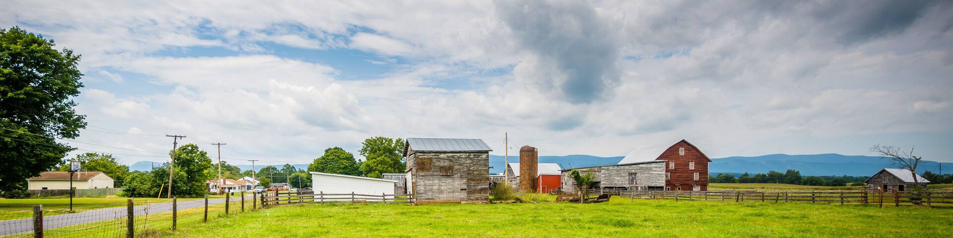 A rural landscape with a green field, a wooden fence, and several barns under a cloudy sky.