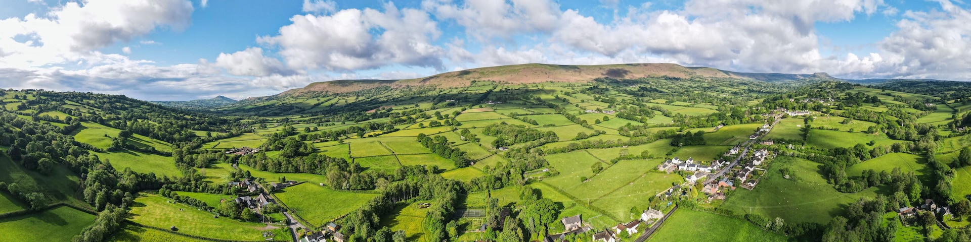 Ein weites Panorama über ein üppiges, grünes Tal mit sanften Hügeln, einem kleinen Dorf und einem markanten Hügel im Hintergrund.