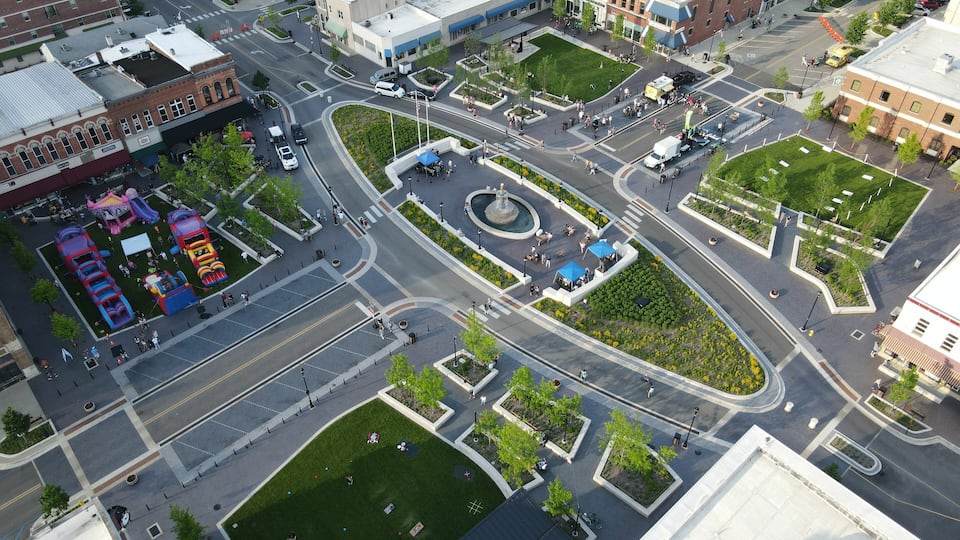 An aerial view of a city intersection with a roundabout, green spaces, and buildings.