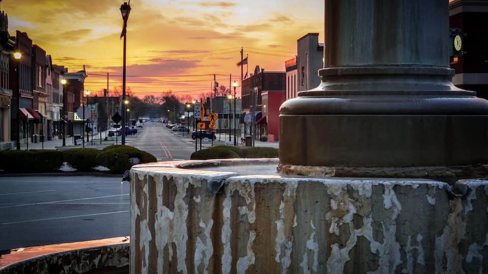 A sunset over a street with buildings, a clock tower, and a weathered monument.