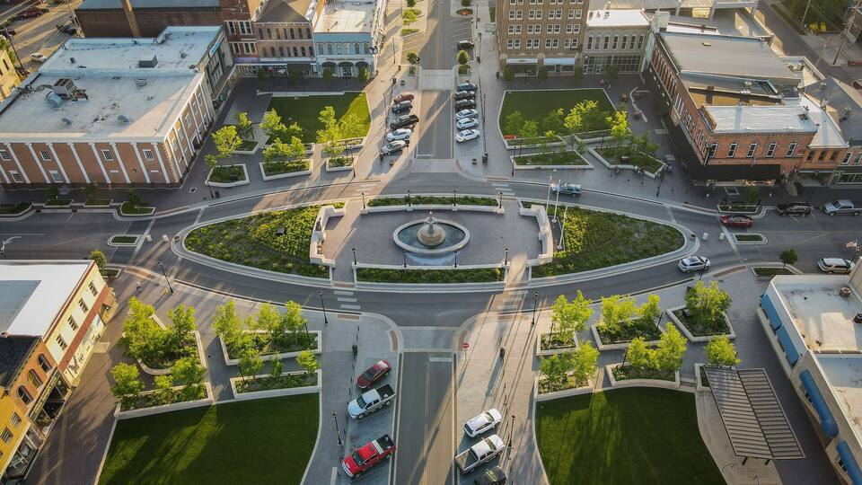 An aerial view of a city intersection with a central roundabout, surrounding buildings, and green spaces.