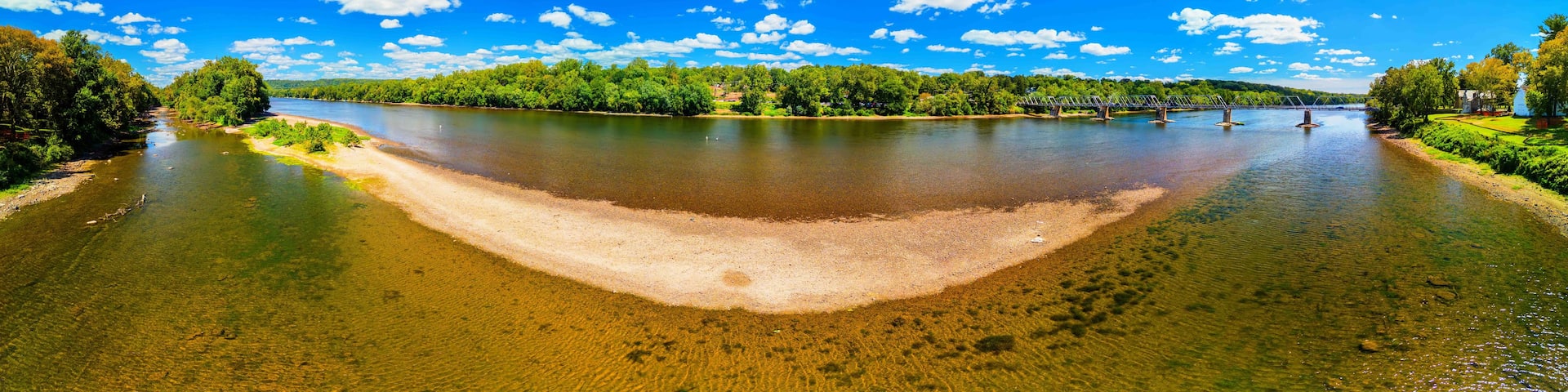 A river with a sandy bank, a bridge, and a forested area under a clear blue sky.
