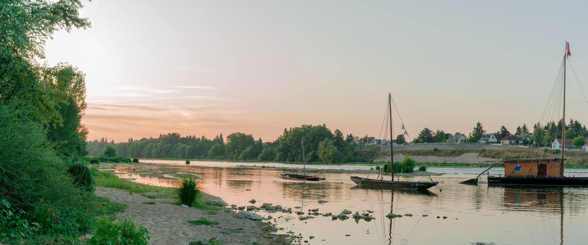 A calm river with boats, a sandy shore, and a forested area in the background.