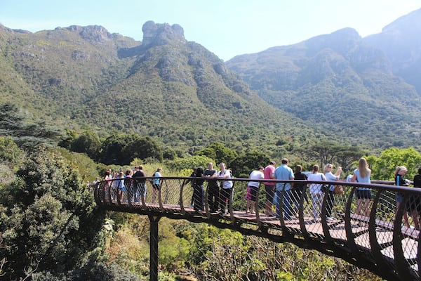 A wooden bridge with people standing on it, overlooking a mountainous landscape with dense greenery.