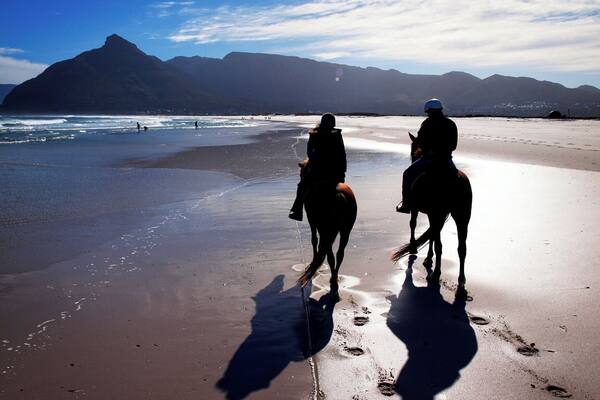 Two people riding horses on a beach with mountains in the background.