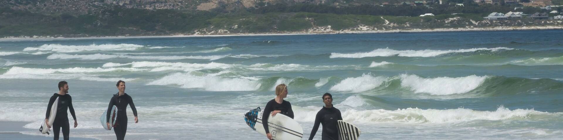 A beach with surfers walking along the shore, waves crashing onto the beach, and a hilly landscape in the background.