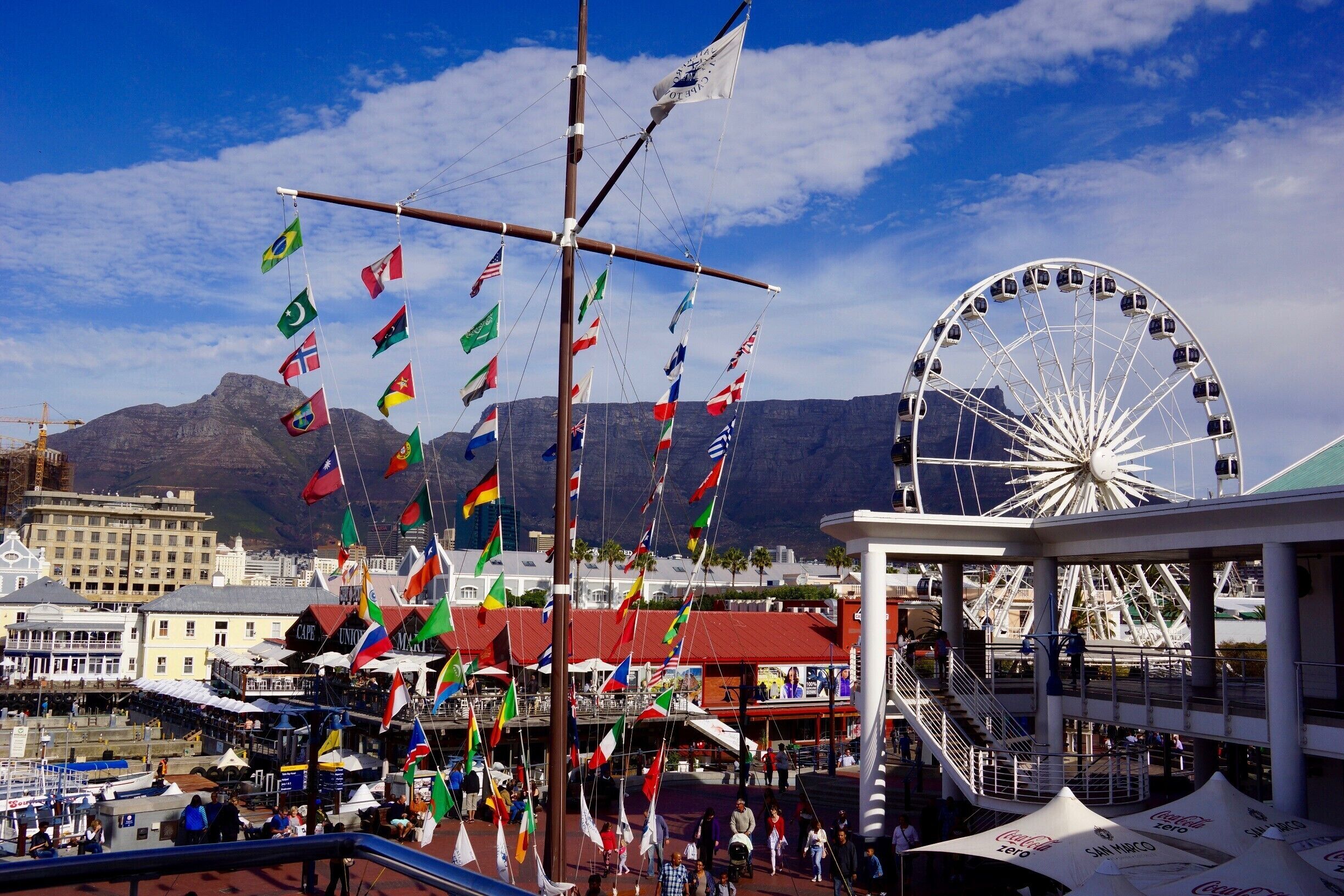 Una feria concurrida con noria, un velero con varias banderas y un telón de fondo montañoso.