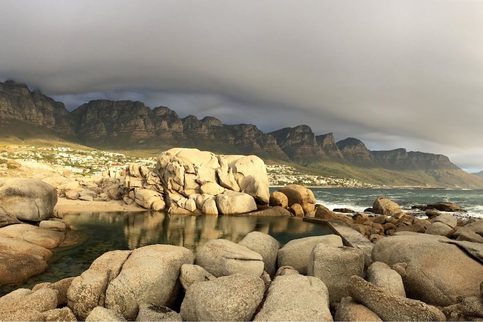 A rocky coastline with large boulders, a small body of water, and a town in the background with mountains in the distance.