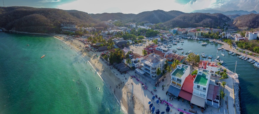 An aerial view of a coastal town with a beach, a marina, and surrounding hills.