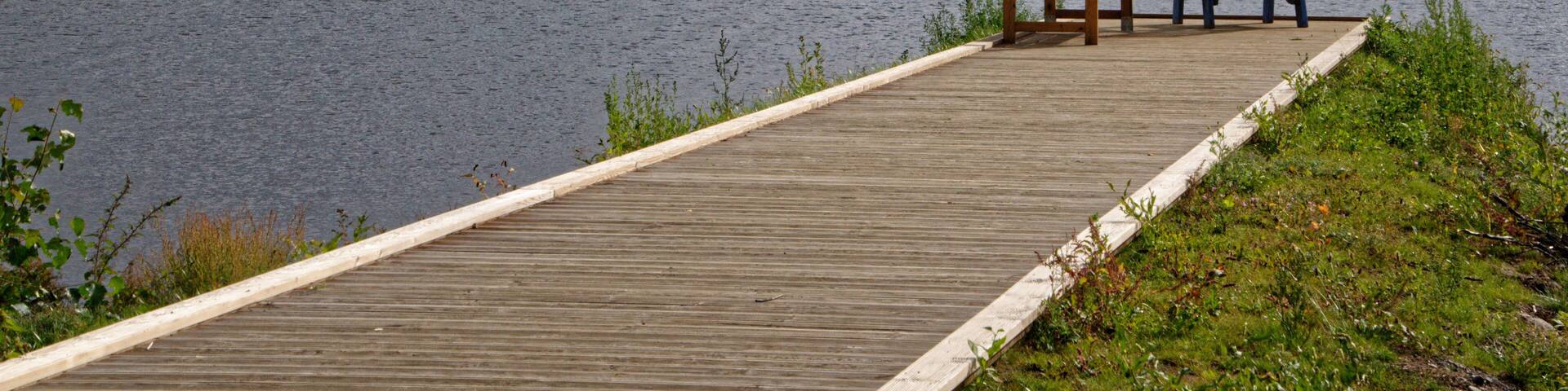 A wooden dock extending into a lake with two chairs and a table, surrounded by dense forest under a cloudy sky.