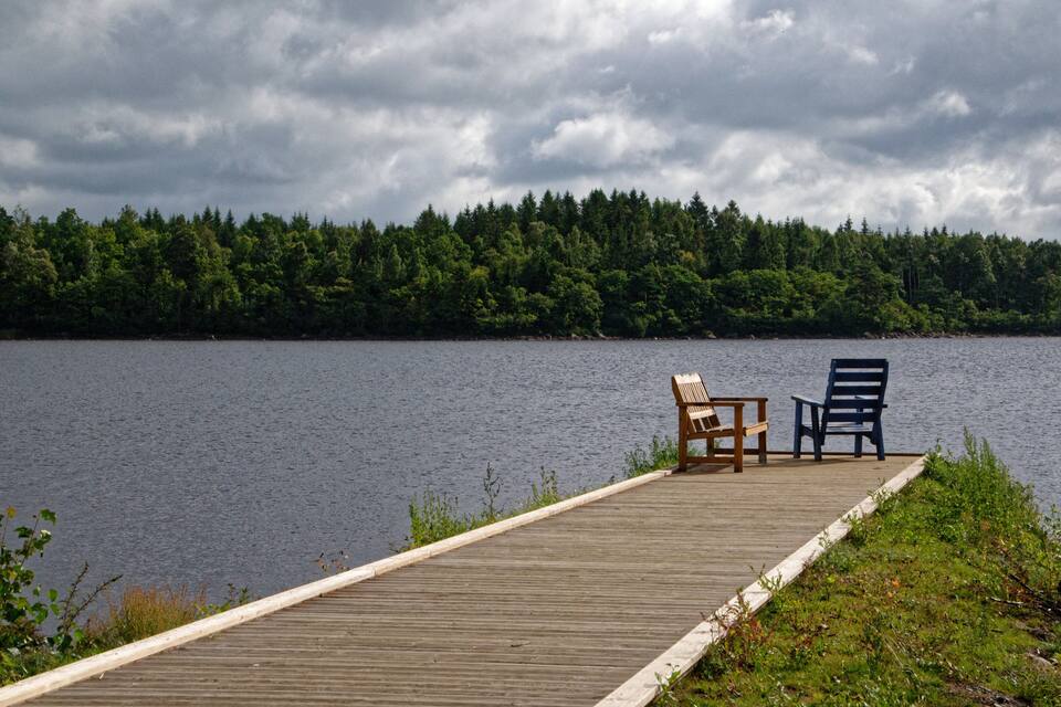 Un molo in legno che si protende in un lago, con due sedie e un tavolo, circondato da una fitta foresta sotto un cielo nuvoloso.