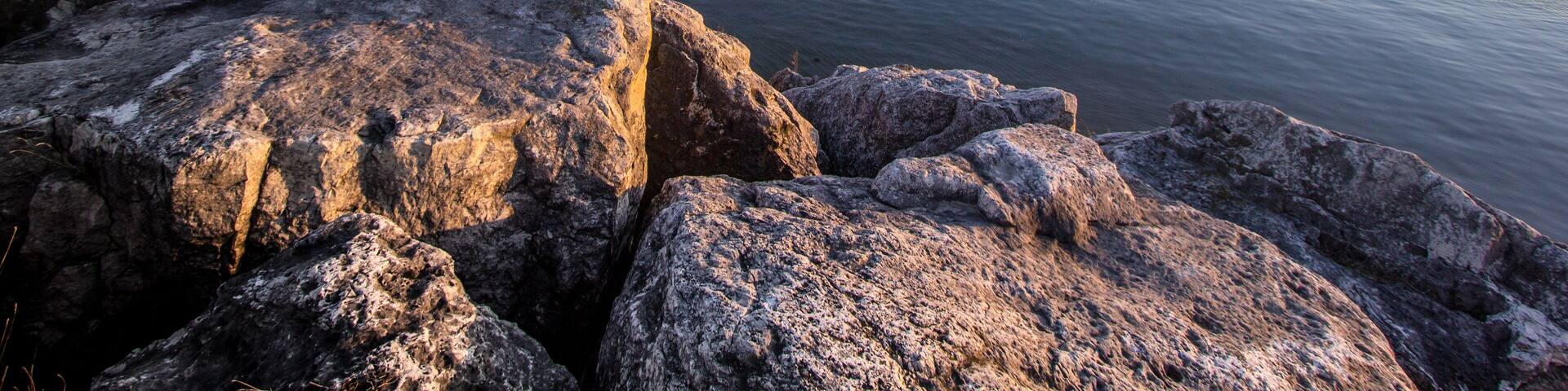 A rocky shoreline with a calm body of water, a distant bridge, and a clear sky.